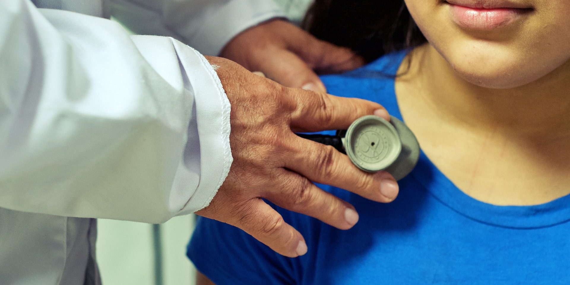 Doctor in white medical coat uses stethoscope on girl wearing blue shirt.