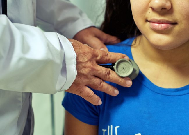 Doctor in white medical coat uses stethoscope on girl wearing blue shirt.