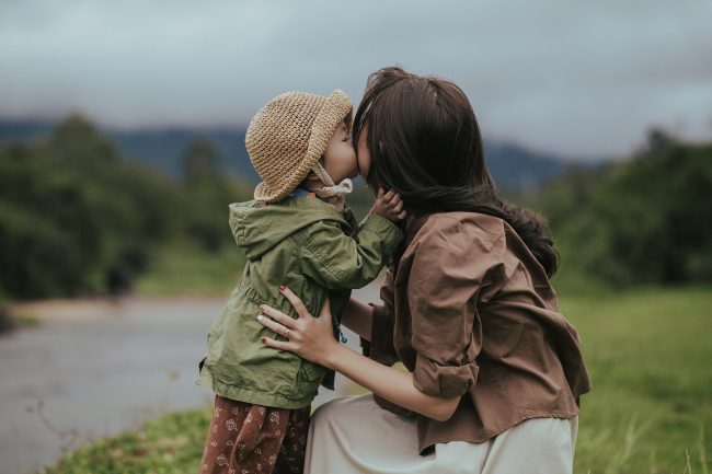 Mother wearing a brown jacket and daughter wearing a green jacket and tan hat embrace with mountain behind them.