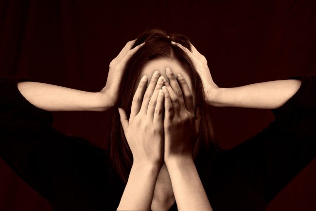 Woman pictured in black shirt with hands over her eyes and another set of hands over her ears.